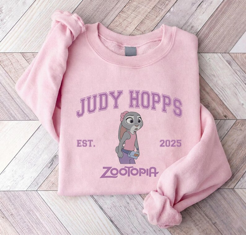 K&ouml;nnte beinhalten: Hellrosa Sweatshirt mit dem Text "JUDY HOPPS" &uuml;ber einer Illustration von Judy Hopps aus Zootopia. Das Sweatshirt enth&auml;lt auch den Text "EST. 2025" und "ZOOTOPIA" in Lila.