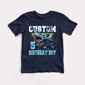 May include: A navy blue t-shirt with a graphic of a blue monster truck with a shark head. The text "CUSTOM 5 BIRTHDAY BOY" is printed on the shirt.