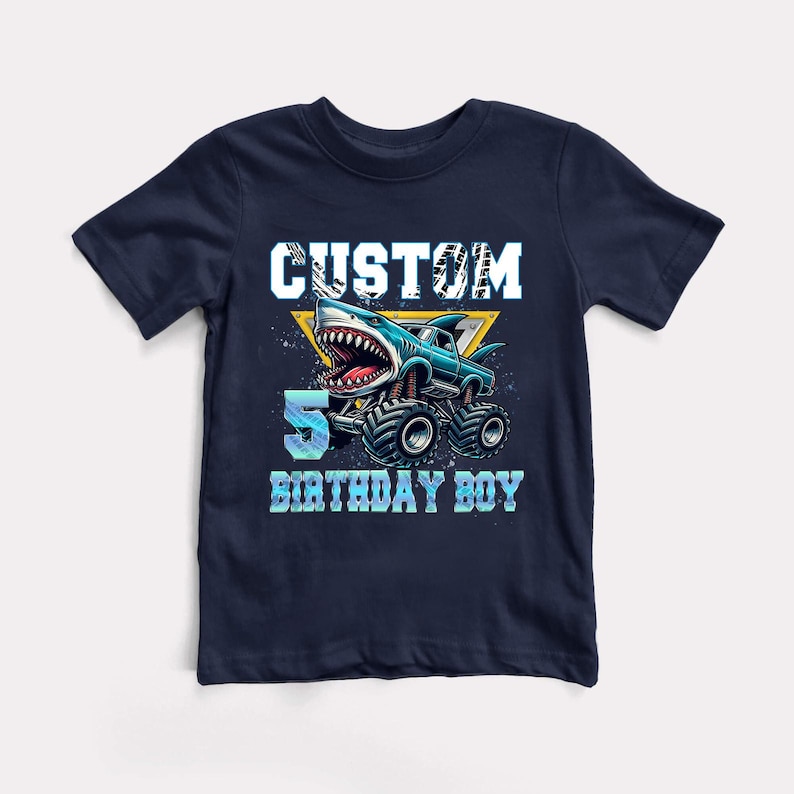 May include: A navy blue t-shirt with a graphic of a blue monster truck with a shark head. The text "CUSTOM 5 BIRTHDAY BOY" is printed on the shirt.