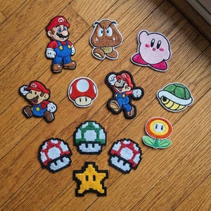 Könnte beinhalten: Eine Sammlung von aufgestickten Aufnähern mit Charakteren und Symbolen aus der Super Mario-Videospielserie. Enthält Mario, Goomba, Kirby, Pilze, einen Stern und eine Blume. Die Aufnäher sind farbenfroh und detailliert, mit verschiedenen Formen und Größen.