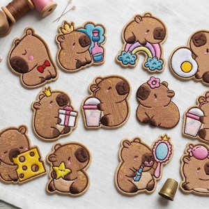 Könnte beinhalten: Eine Sammlung von aufgestickten Aufnähern mit Cartoon-Capybaras in verschiedenen Posen. Einige Capybaras halten Gegenstände wie ein Geschenk, ein Getränk oder einen Spiegel. Die Aufnäher sind braun mit detaillierten Stickereien und farbenfrohen Akzenten.
