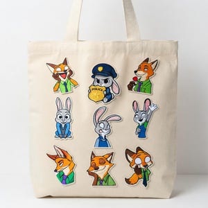 May include: A beige tote bag featuring embroidered patches of cartoon animals. The patches include a fox, a bunny, and other characters in various poses. The patches are in shades of orange, blue, green, and white.
