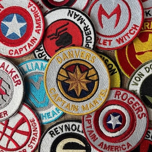 May include: A collection of embroidered patches featuring superhero emblems and names. The patches are round with various colors, including red, blue, and gold. Text includes "Captain America," "Spider-Man," and "Captain Marvel."