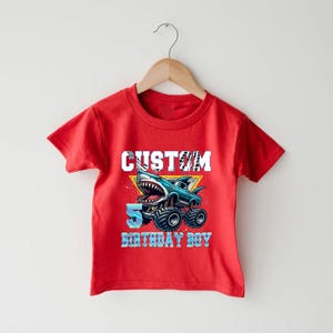 May include: Red short-sleeved t-shirt with a graphic of a monster truck with a shark fin and the text "CUSTOM 5 BIRTHDAY BOY".