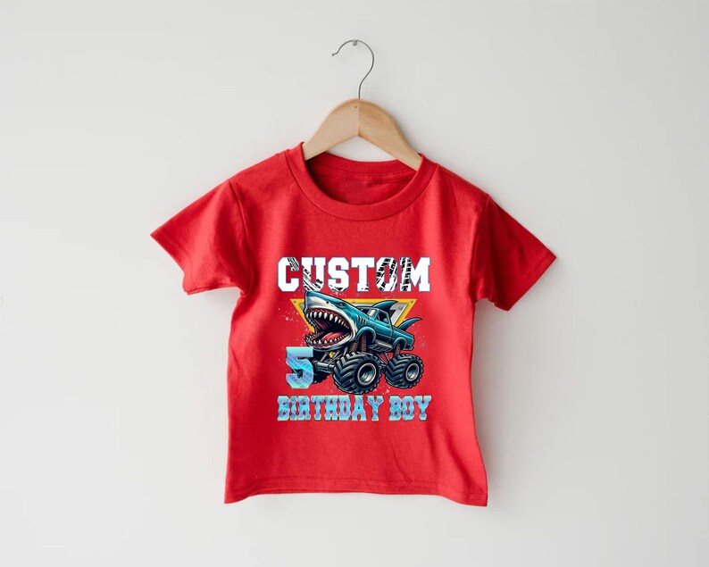 May include: Red short-sleeved t-shirt with a graphic of a monster truck with a shark fin and the text "CUSTOM 5 BIRTHDAY BOY".