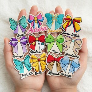 Könnte beinhalten: Eine Sammlung gestickter Schleifenaufnäher in verschiedenen Farben, jeweils mit einem Charakternamen wie Ariel, Aurora und Jasmine. Die Aufnäher weisen detaillierte Stickereien auf und werden in offenen Händen gehalten.
