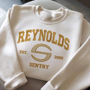 May include: A cream-coloured sweatshirt featuring gold embroidered text. The text reads "REYNOLDS" above a stylized "S" and "SENTRY" below. The sweatshirt also has "EST. 2000". The cuffs and collar are ribbed.