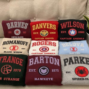 May include: Nine folded sweatshirts with embroidered text and graphics. The sweatshirts are in various colours, including maroon, red, black, cream, white, light blue, dark green, and grey. Each sweatshirt features a different name and year, such as "BARNES" and "1917".