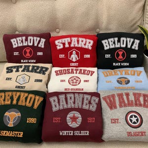 May include: Ten different colored sweatshirts with embroidered logos and text. The sweatshirts are arranged in two rows of five. The text on the sweatshirts includes the names "BELOVA", "STARR", "SHOSATAKOV", "DREYKOV", "BARNES", and "WALKER".