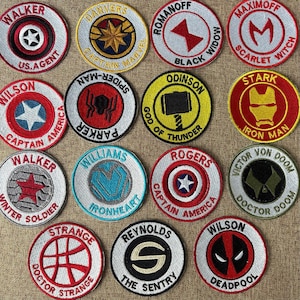 Könnte beinhalten: Eine Sammlung gestickter Aufnäher mit Superhelden-Logos und -Namen. Die runden Aufnäher sind in verschiedenen Farben wie Rot, Blau, Gelb und Weiß gehalten. Jeder Aufnäher hat ein einzigartiges Design und Text.