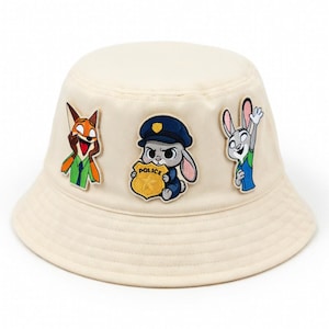 May include: Cream-colored bucket hat featuring embroidered patches of cartoon characters. The patches include a fox in a green and orange outfit, a bunny in a police uniform, and a bunny waving. The hat is a casual accessory.