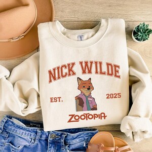 K&ouml;nnte beinhalten: Cremefarbenes Sweatshirt mit dem Text "NICK WILDE" in roten, bogenf&ouml;rmigen Buchstaben &uuml;ber einer Illustration eines Fuchses. Darunter stehen die Worte "EST. 2025" und "ZOOTOPIA" in Rot. Das Sweatshirt wird mit blauen Jeans und einem beigen Hut kombiniert.