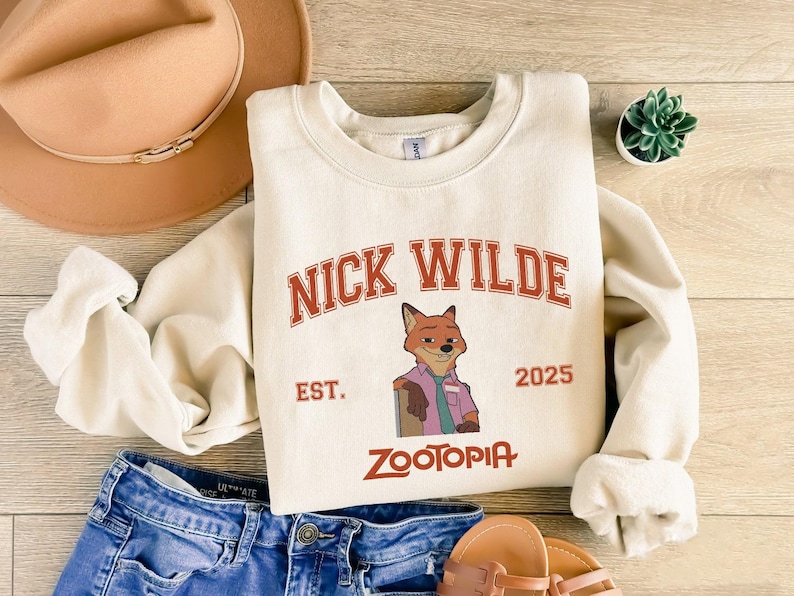 K&ouml;nnte beinhalten: Cremefarbenes Sweatshirt mit dem Text "NICK WILDE" in roten, bogenf&ouml;rmigen Buchstaben &uuml;ber einer Illustration eines Fuchses. Darunter stehen die Worte "EST. 2025" und "ZOOTOPIA" in Rot. Das Sweatshirt wird mit blauen Jeans und einem beigen Hut kombiniert.