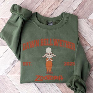 K&ouml;nnte beinhalten: Gr&uuml;nes Sweatshirt mit der Figur Dawn Bellwether aus Zootopia. Das Design umfasst den Text "DAWN BELLWETHER", "EST. 2025" und "ZOOTOPIA" in Orange. Das Sweatshirt hat einen Rundhalsausschnitt und lange &Auml;rmel.