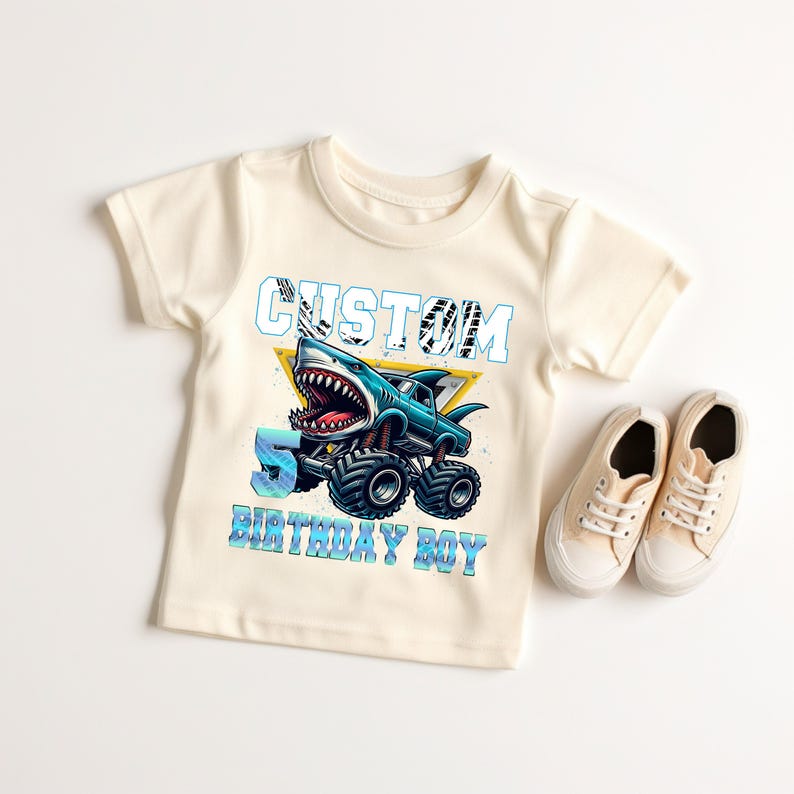 May include: A light beige t-shirt with a graphic of a blue monster truck with a shark head. The truck has a yellow cab and black wheels. The text "CUSTOM" is above the truck and "5 BIRTHDAY BOY" is below the truck.