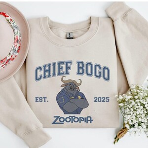 K&ouml;nnte beinhalten: Beiges Sweatshirt mit dem Text "CHIEF BOGO" in blauen Buchstaben und einer Illustration eines B&uuml;ffelcharakters. Der Text "EST. 2025" und "ZOOTOPIA" sind ebenfalls auf dem Sweatshirt aufgedruckt. Ein Hut und Blumen sind ebenfalls im Bild.
