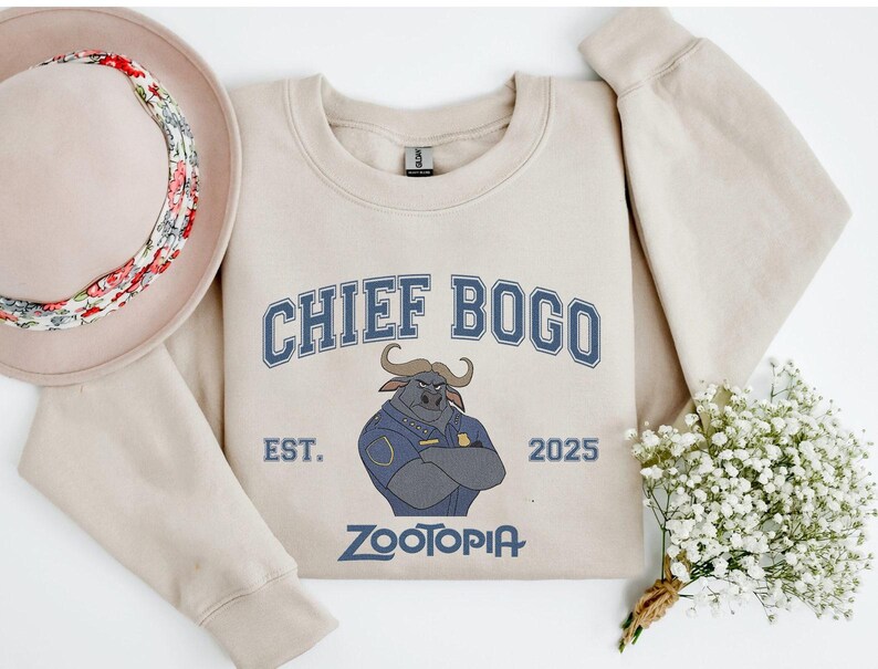 K&ouml;nnte beinhalten: Beiges Sweatshirt mit dem Text "CHIEF BOGO" in blauen Buchstaben und einer Illustration eines B&uuml;ffelcharakters. Der Text "EST. 2025" und "ZOOTOPIA" sind ebenfalls auf dem Sweatshirt aufgedruckt. Ein Hut und Blumen sind ebenfalls im Bild.