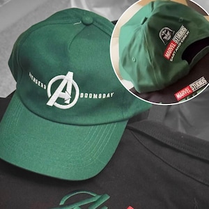 May include: Green baseball cap with white embroidered "Avengers Doomsday" logo on the front. The cap also features a small patch with "Marvel Studios Cast and Crew" text. The cap is made of a soft material.