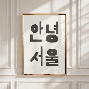 Puede incluir: Un cuadro decorativo rectangular vertical en un marco dorado fino que presenta un texto coreano negro en negrita que dice "안녕 서울" sobre un fondo blanco liso. La tipografía es gruesa y sin remates, centrada dentro del marco, creando un contraste.