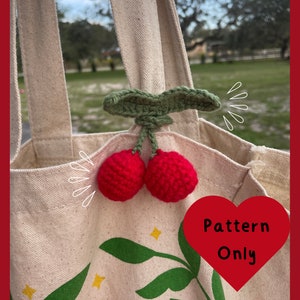 Puede incluir: Una bolsa de lona blanca con un amuleto de cereza de ganchillo verde y rojo. La bolsa tiene un estampado floral verde y amarillo y el texto "Pattern Only" en un corazón rojo.