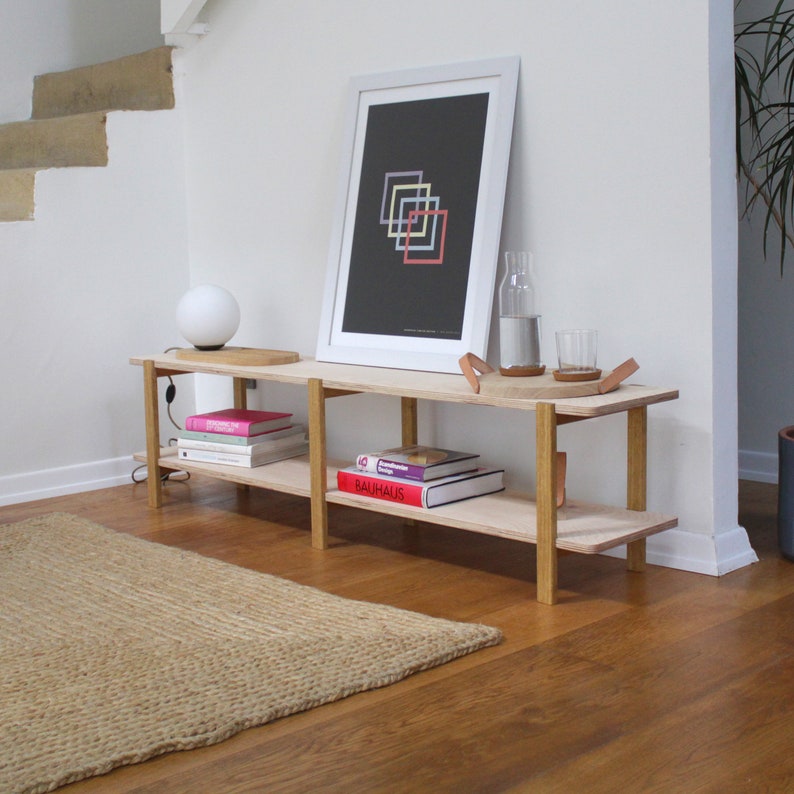May include: A light wood shelf unit with two shelves, featuring a white framed print with a geometric design, a lamp, and books. The shelf unit is against a white wall and sits on a light brown wood floor.