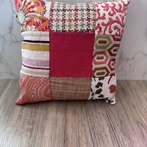 Puede incluir: Un cojín cuadrado de patchwork con una variedad de telas estampadas en tonos rojos, naranjas, rosas y beige. El cojín presenta diseños florales, geométricos y a rayas, creando un acento decorativo.
