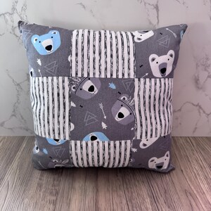 Puede incluir: Cojín decorativo cuadrado con diseño de patchwork. El cojín presenta tela gris con rayas blancas y caras de oso en azul y gris. El cojín mide aproximadamente 30 cm cuadrados.