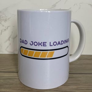 Peut inclure: Mug en céramique blanche avec l'inscription "DAD JOKE LOADING" en violet, au-dessus d'une barre de chargement jaune et noire. La tasse a une anse incurvée. Fond clair.