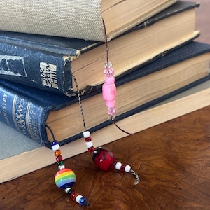 May include: A stack of old books with decorative beaded bookmarks. One bookmark features a rainbow-colored bead, while the other has a red bead with white accents. The books have aged covers in shades of blue, gray, and tan.