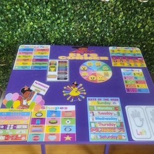 May include: Purple learning table with various educational posters and activities for children. The posters include the alphabet, numbers, days of the week, shapes, money, and more. The table is decorated with a "Kid's Corner" sign and a colorful wheel with different emotions.