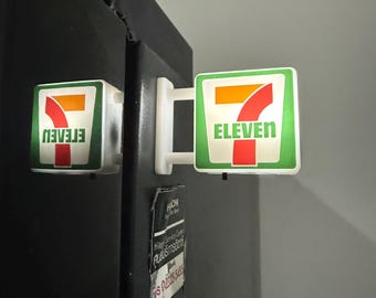 May include: A miniature 7-Eleven sign with a white frame, green border, and the orange number 7 with the word ELEVEN in green text. The sign is mounted on a black surface, with a reflection visible on the side of the black object.