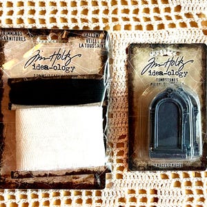 May include: Two Tim Holtz idea-ology packages. The left package contains one spool of black ribbon and one spool of white ribbon. The right package contains two dark grey, arched tombstone-shaped embellishments with a decorative top detail.