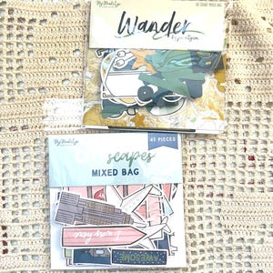 May include: Two packages of paper ephemera. The top package reads "Wander" and contains travel-themed die-cuts. The bottom package reads "scapes MIXED BAG" and includes various die-cuts with the word "AWESOME". Both packages are on a cream-colored crocheted surface.