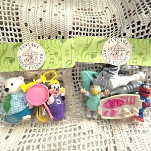May include: Two clear plastic bags containing assorted miniature plastic toys, each topped with a green cardboard header labelled Vintage Paper Trinkets. The left bag holds a polar bear, a pink pan, and a purple figure. The right bag contains a small doll, a red character, and various white and pink plastic accessories.