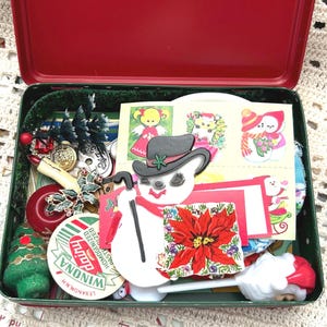 May include: A red metal tin filled with Christmas-themed decorations and ephemera. Items include snowman cutouts, stickers, ornaments, and a green felt Christmas tree. The tin is open, revealing its contents.