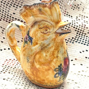 May include: A ceramic pitcher with a rooster design. The pitcher is primarily yellow with brown accents and floral details. The rooster's head forms the spout, and a handle is on the side. The pitcher is hand-painted.