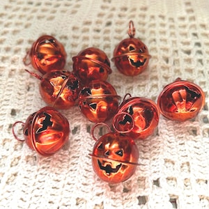 May include: A group of orange-red metal pumpkin-shaped bells with carved faces and hanging loops. They are arranged on a white, textured surface. These decorative bells are ideal for Halloween and add a festive touch to any setting.