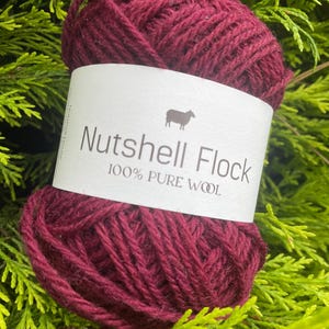 Puede incluir: Un ovillo de lana de color rojo burdeos oscuro con una etiqueta blanca que dice "Nutshell Flock 100% Pure Wool" y una peque&ntilde;a imagen de una oveja.