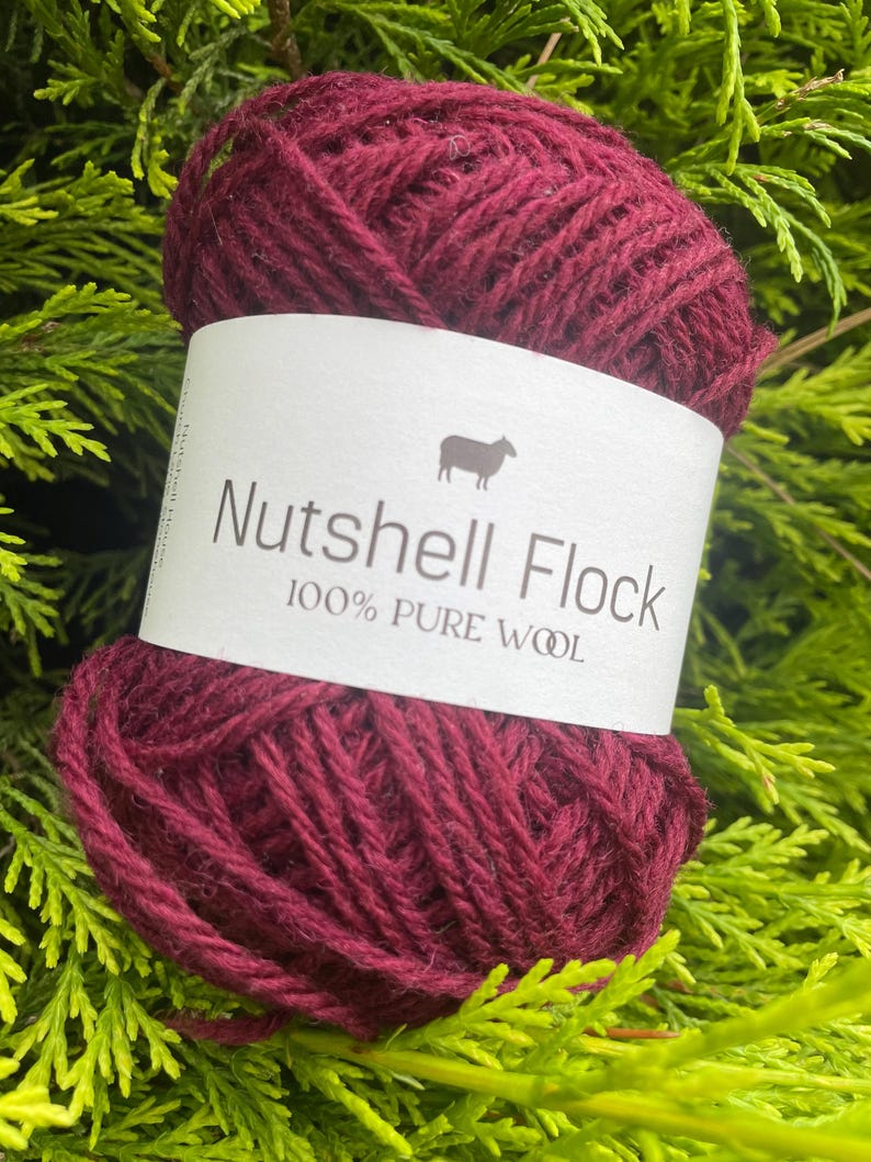 Puede incluir: Un ovillo de lana de color rojo burdeos oscuro con una etiqueta blanca que dice "Nutshell Flock 100% Pure Wool" y una peque&ntilde;a imagen de una oveja.
