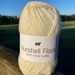 Puede incluir: Un ovillo de lana de color crema con la etiqueta "Nutshell Flock 100% Lana Pura" y una peque&ntilde;a ilustraci&oacute;n de una oveja.