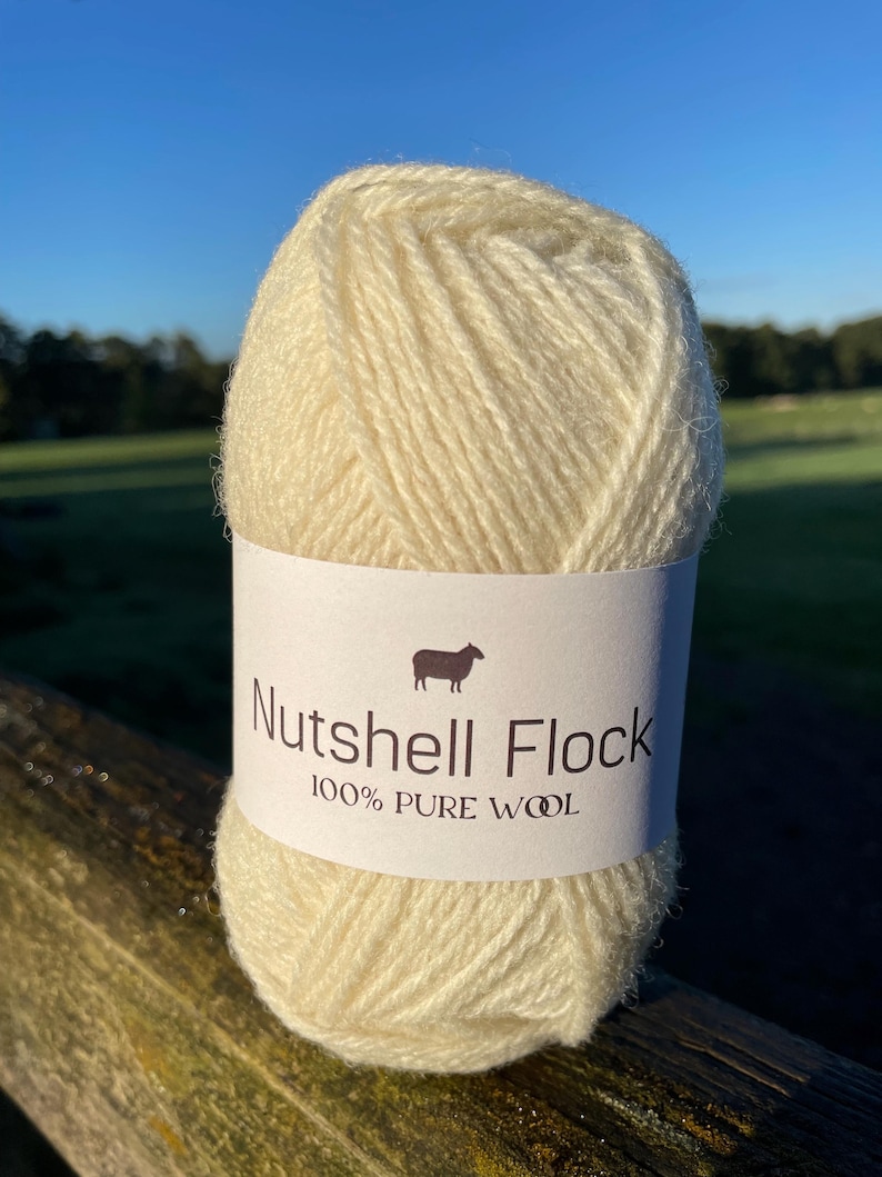 Puede incluir: Un ovillo de lana de color crema con la etiqueta "Nutshell Flock 100% Lana Pura" y una peque&ntilde;a ilustraci&oacute;n de una oveja.