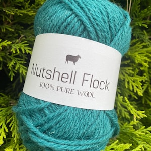 Puede incluir: Una madeja de lana de color turquesa con la etiqueta "Nutshell Flock 100% Lana Pura" y un icono de oveja.