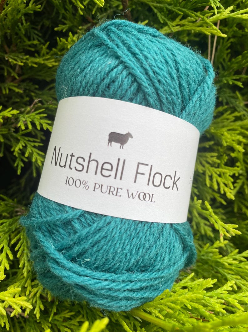 Puede incluir: Una madeja de lana de color turquesa con la etiqueta "Nutshell Flock 100% Lana Pura" y un icono de oveja.