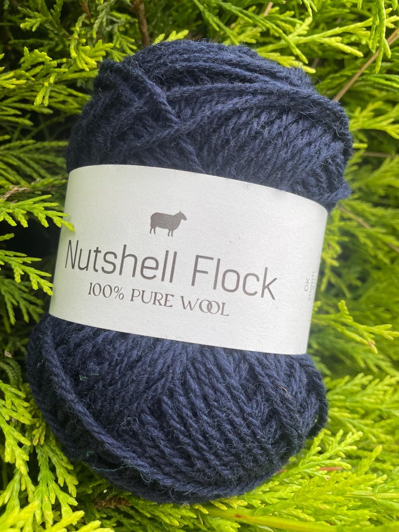 Puede incluir: Una madeja de lana azul oscuro etiquetada "Nutshell Flock 100% Lana Pura" con una peque&ntilde;a ilustraci&oacute;n de oveja.