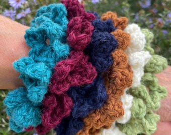 Puede incluir: Una colección de coleteros de ganchillo hechos a mano en varios colores, incluyendo turquesa, burdeos, azul marino, marrón, blanco y verde. Estos accesorios para el cabello de tela están dispuestos en un patrón circular, mostrando la textura y las variaciones de color.