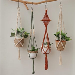 Op de afbeelding: Vier macramé plantenhangers met houten kralen en kwastjes die aan een tak hangen. Elke hanger heeft een kleine plant in een pot.