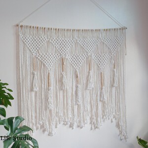 May include: A white macrame wall hanging with wooden beads and tassels. The hanging is made of cotton cord and has a geometric pattern.