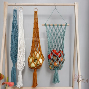 May include: Macrame hanging fruit baskets in teal, white, and rust colors, displayed on a wooden rack. One basket holds onions, and another holds tomatoes and onions. Each basket has a tassel.