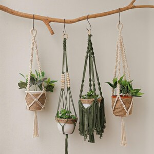 Può includere: Quattro portavasi in macramè con stili e colori diversi. I portavasi sono realizzati in fibre naturali e hanno perline di legno. Sono appesi a un ramo su una parete bianca.