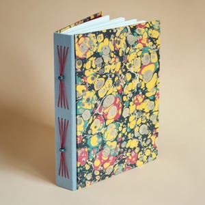 May include: A handmade journal with a marbled cover featuring yellow, red, and teal patterns on a black background. The spine is light blue with red stitching and teal beads. The pages are white.